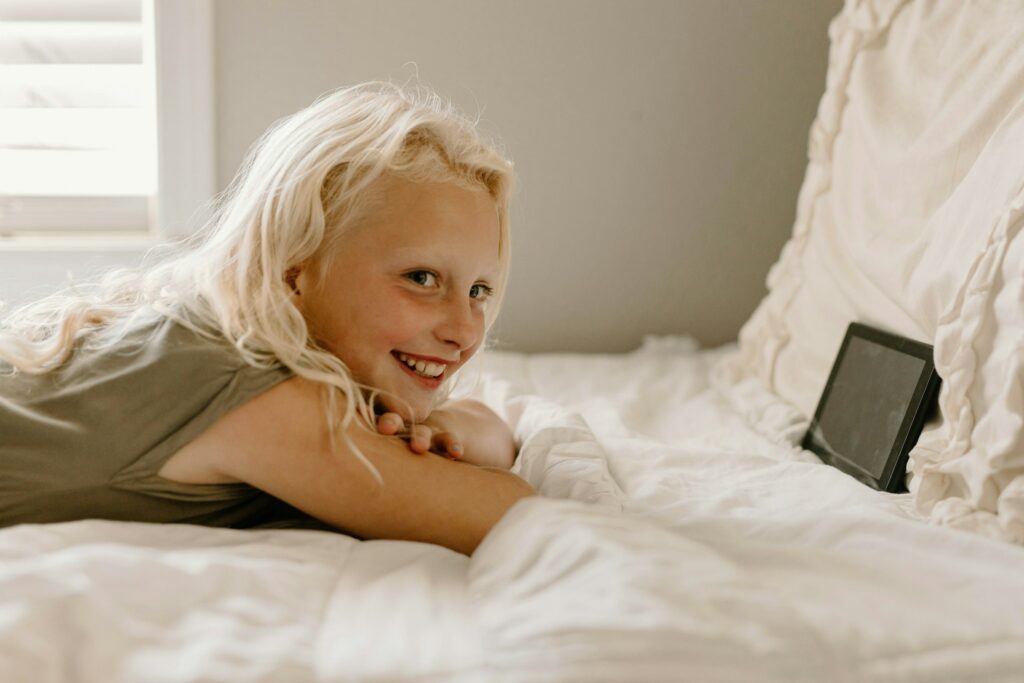 A young girl lying on a bed, smiling with a tablet resting beside her, protected by electrical safety measures for children.